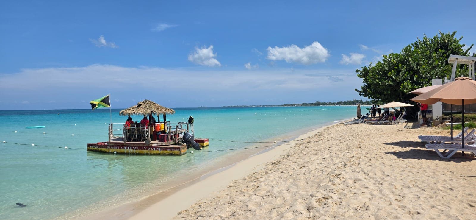 Seven Mile Beach at midday, turquoise shallows, a boat under the Jamaican flag moored off white sand